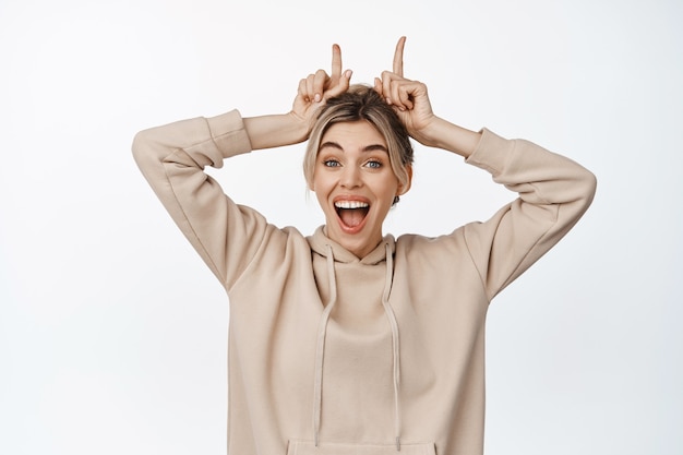 Foto grátis mulher jovem animada parece feliz e surpresa, mostrando os dedos apontando na cabeça, gesto de chifres de touro, de pé no branco.