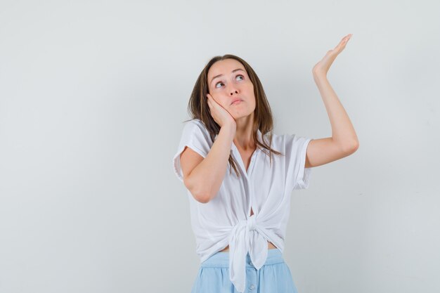 Mulher jovem apoiando a bochecha na palma da mão e levantando a outra mão no ar em uma blusa branca e saia azul claro e parecendo pensativa