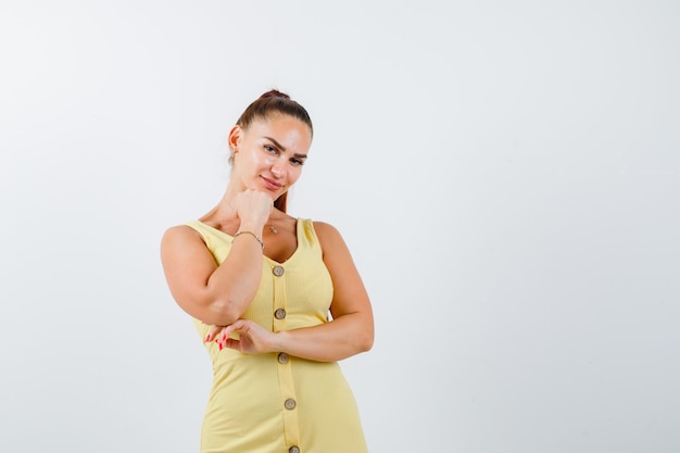 Mulher jovem, apoiando o queixo no punho em um vestido amarelo e parecendo sensata. vista frontal.