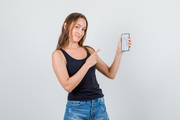 Foto grátis mulher jovem apontando o dedo para o smartphone em camiseta, shorts e parecendo alegre