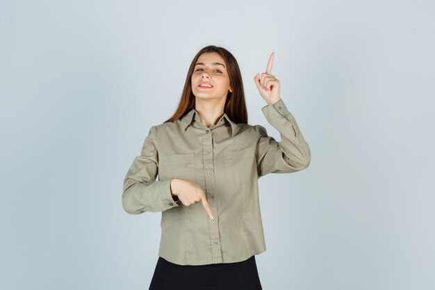 Mulher jovem apontando os dedos para cima e para baixo na camisa, saia e parecendo orgulhosa