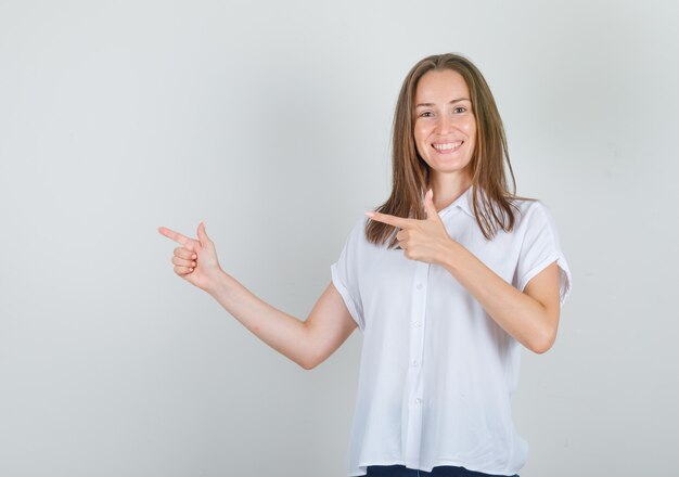 Mulher jovem apontando os dedos para o lado em uma camiseta branca e parecendo alegre