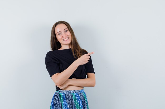 Mulher jovem apontando para a direita com o dedo indicador em uma camiseta preta e saia azul e parece atraente