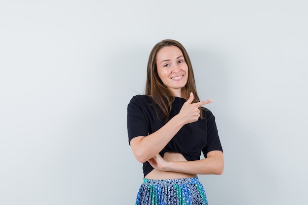 Mulher jovem apontando para a direita com o dedo indicador em uma camiseta preta e saia azul e parecendo feliz