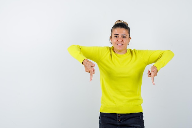 Mulher jovem apontando para baixo com os dedos indicadores em um suéter amarelo e calça preta e parecendo preocupada