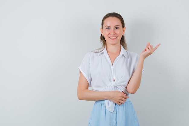 Mulher jovem apontando para o canto superior direito com blusa, saia e parecendo alegre