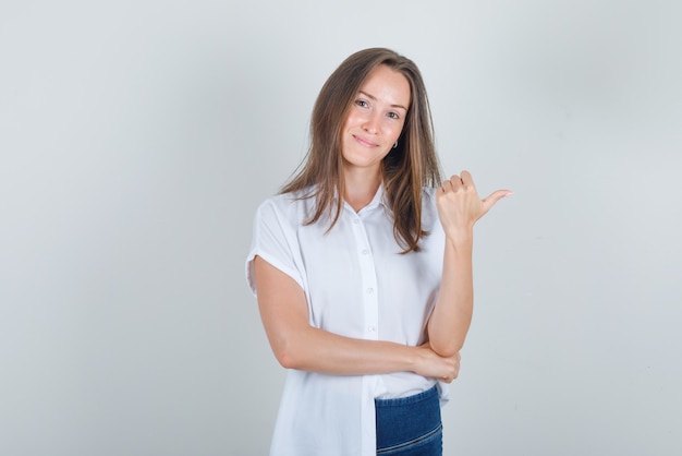 Mulher jovem apontando para o lado com o polegar para cima em uma camiseta, jeans e parecendo alegre