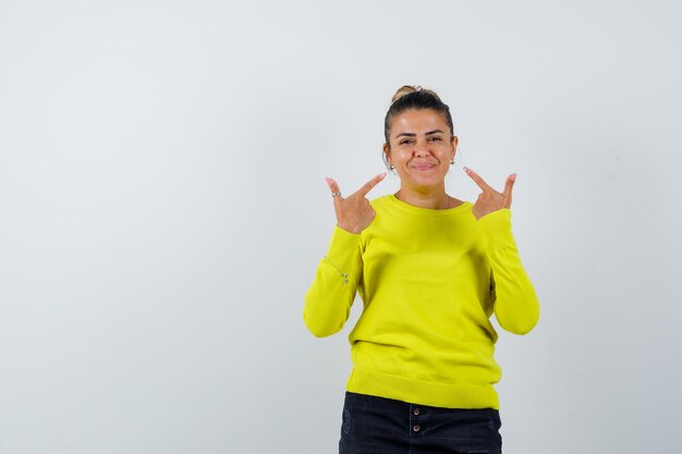 Mulher jovem apontando para si mesma com um suéter amarelo e calça preta e parecendo feliz