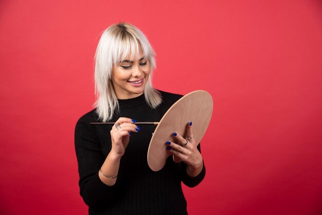 Mulher jovem artista segurando materiais de arte em uma parede vermelha.