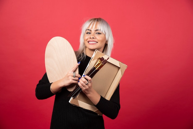 Mulher jovem artista segurando materiais de arte em uma parede vermelha.