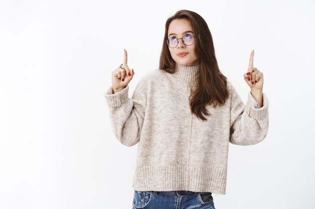 Mulher jovem atraente e elegante de óculos e suéter aconchegante e aconchegante franzindo os lábios coquete, sorrindo, olhando e apontando para cima com interesse