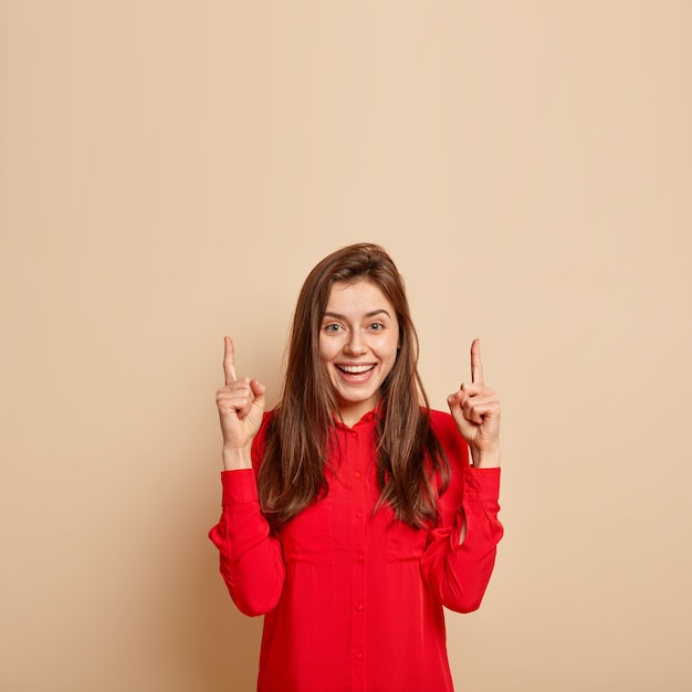 Mulher jovem bonita e encantada aponta para cima, parece impressionada e fascinada, vestida com camisa da moda vermelha, demonstra item contra parede bege, mostra espaço em branco para sua promoção