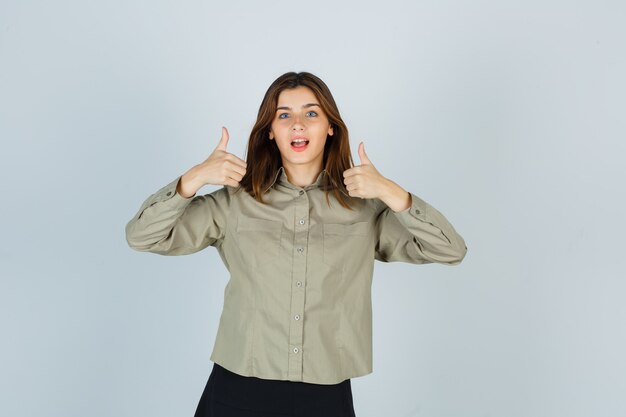 Mulher jovem bonita mostrando dois polegares para cima na camisa, saia e olhando maravilhado, vista frontal.