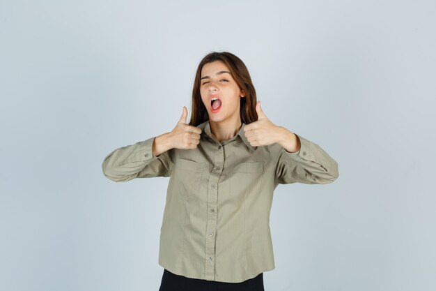 Mulher jovem bonita na camisa mostrando os polegares duplos enquanto pisca e parece confiante, vista frontal.