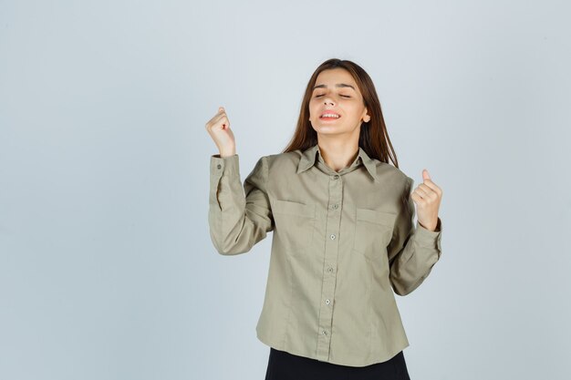 Mulher jovem bonita na camisa, saia, mostrando o gesto do vencedor e olhando feliz, vista frontal.