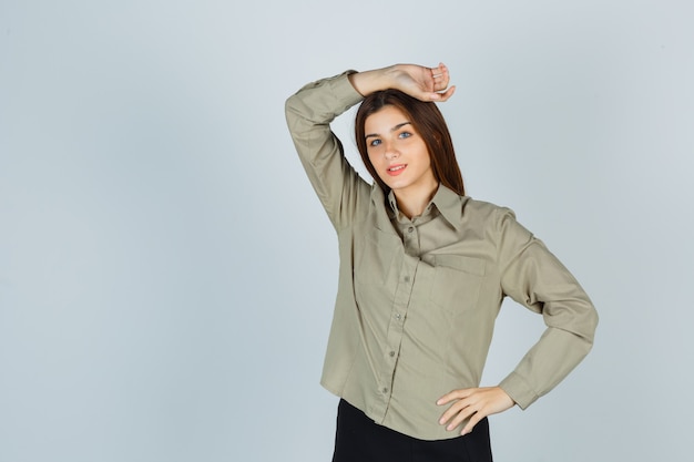 Mulher jovem bonita posando com a mão na cabeça na camisa, saia e olhando alegre. vista frontal.