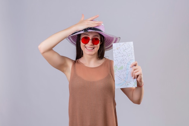 Mulher jovem bonita viajante com chapéu de verão, óculos de sol vermelhos segurando bilhetes aéreos, sorrindo com cara feliz sobre parede branca