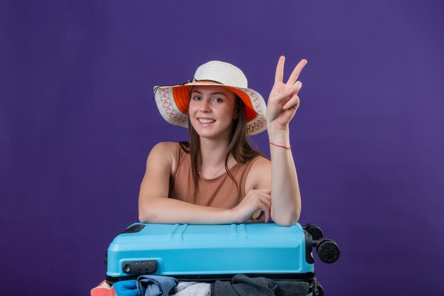 Mulher jovem bonita viajante no chapéu de verão com mala cheia de roupas positivas e felizes sorrindo alegremente otimistas mostrando sinal de vitória ou número dois sobre parede roxa