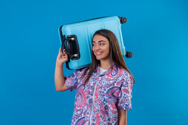 Foto grátis mulher jovem bonita viajante segurando a mala azul, olhando de lado com cara feliz sorrindo alegremente sobre parede azul