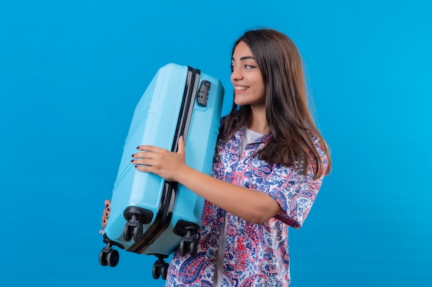 Mulher jovem bonita viajante segurando a mala azul, olhando de lado positivo e feliz sorrindo alegremente sobre parede azul