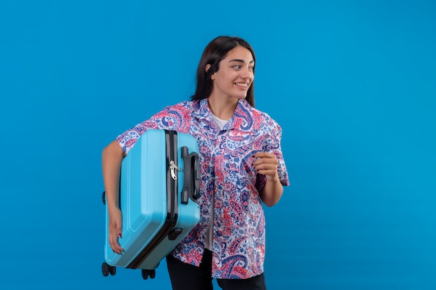 Mulher jovem bonita viajante segurando mala olhando confiante, positivo e feliz, sorrindo alegremente, pronto para viajar sobre parede azul