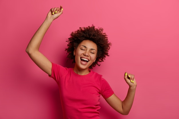 Mulher jovem cacheada otimista e despreocupada levanta os braços e dança ao som da música incrível favorita, se move com o ritmo da música, usa uma camiseta casual, isolada sobre a parede rosa, esquece todos os problemas