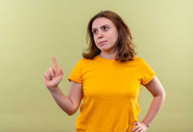 Mulher jovem casual apontando para cima com a mão na cintura em uma parede verde isolada com espaço de cópia