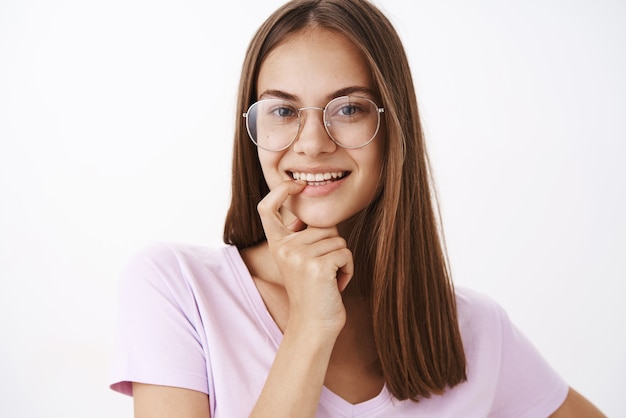 Mulher jovem charmosa, glamourosa e confiante de óculos, segurando o dedo no lábio inferior e sorrindo com desejo