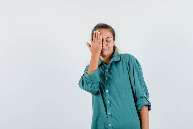 Mulher jovem cobrindo o olho direito com a mão na camisa azul e parecendo feliz