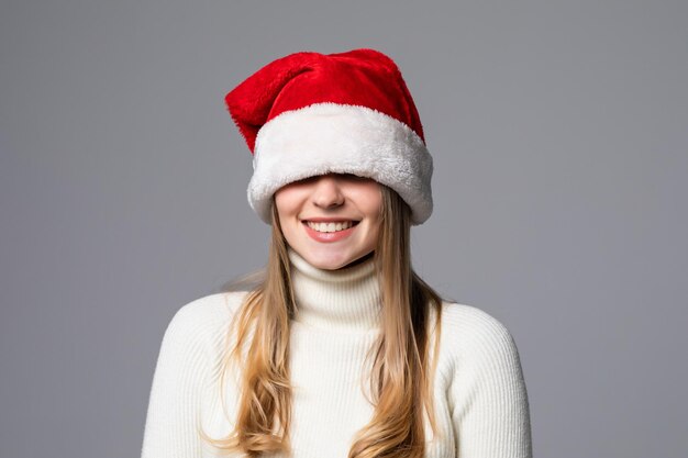 Mulher jovem cobrindo os olhos com um chapéu de Papai Noel isolado na parede cinza