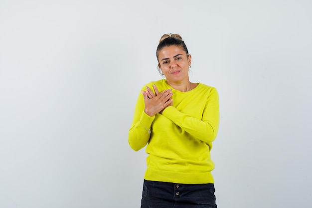 Mulher jovem com as mãos no peito, suéter amarelo e calça preta, parecendo séria