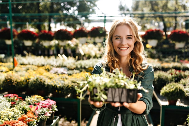 Mulher jovem com belos cabelos loiros e sorriso gentil, vestida com um manto verde com cinto está trabalhando em estufa