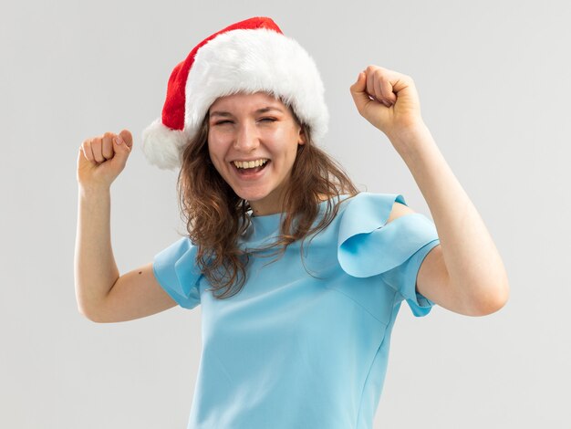 Mulher jovem com blusa azul e chapéu de Papai Noel cerrando os punhos feliz e animada