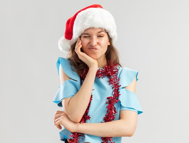 Mulher jovem com blusa azul e chapéu de Papai Noel com enfeites em volta do pescoço e expressão cética