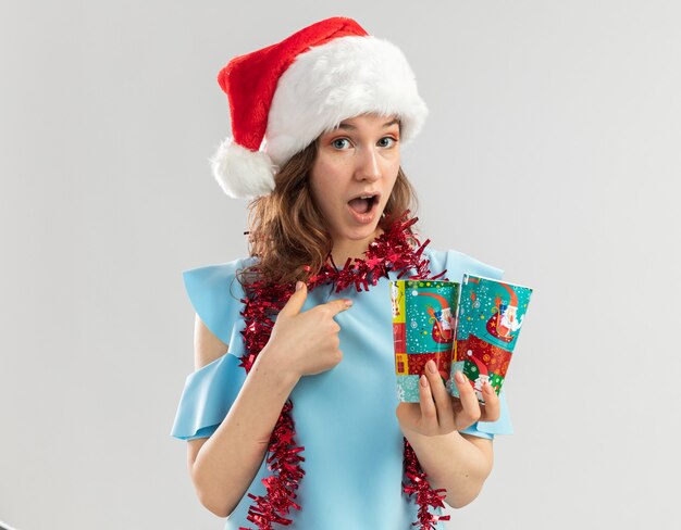 Mulher jovem com blusa azul e chapéu de Papai Noel com enfeites em volta do pescoço segurando copos de papel coloridos parecendo surpresa apontando para si mesma
