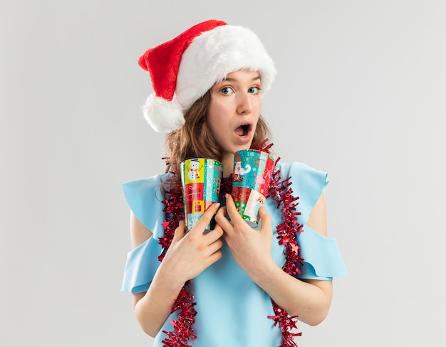 Mulher jovem com blusa azul e chapéu de Papai Noel com enfeites em volta do pescoço segurando copos de papel coloridos parecendo surpresa