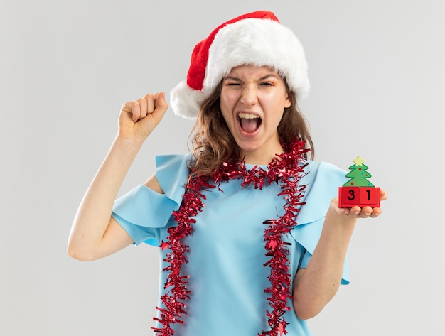 Mulher jovem com blusa azul e chapéu de Papai Noel com enfeites em volta do pescoço segurando cubos de brinquedo com data do ano feliz ney cerrando os punhos gritando celebrando a loucura feliz