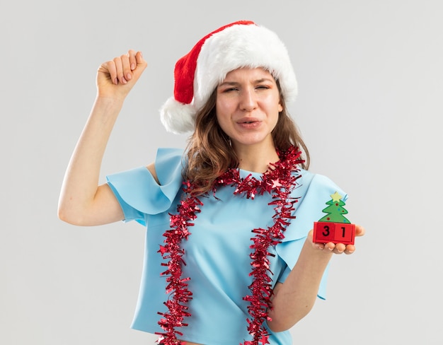 Mulher jovem com blusa azul e chapéu de Papai Noel com enfeites em volta do pescoço segurando cubos de brinquedo com data feliz ano ney cerrando o punho feliz e animada