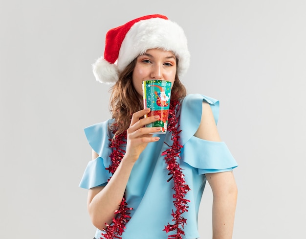 Mulher jovem com blusa azul e chapéu de Papai Noel com enfeites no pescoço segurando um copo de papel colorido, parecendo feliz e positiva