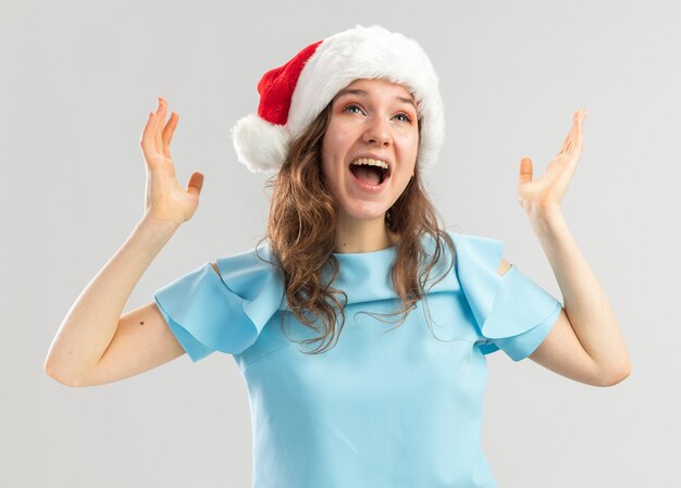 Mulher jovem com blusa azul e chapéu de papai noel gritando com os braços erguidos loucamente feliz e animada