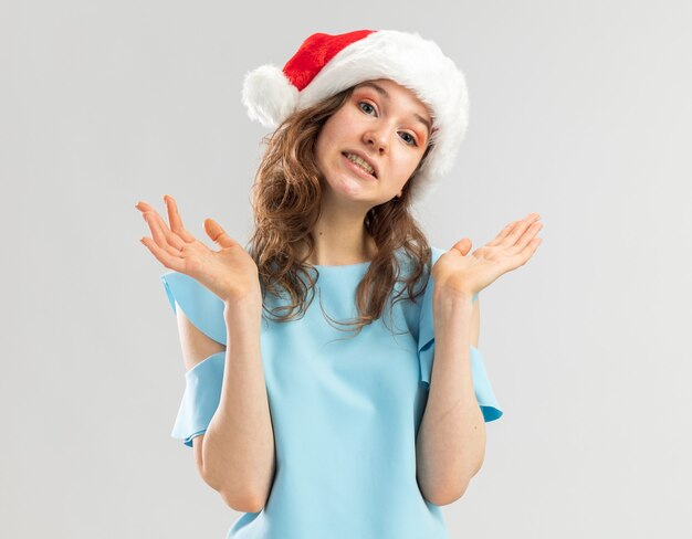 Mulher jovem com blusa azul e chapéu de Papai Noel, parecendo confusa, levantando os braços