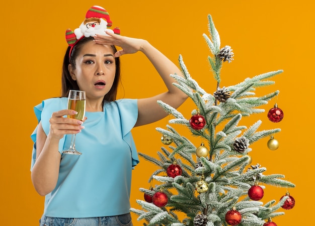 Mulher jovem com blusa azul, usando um aro de natal engraçado na cabeça, segurando uma taça de champanhe, olhando para longe sendo surpreendida ao lado de uma árvore de Natal sobre fundo laranja