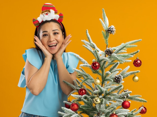 Mulher jovem com blusa azul, usando uma borda de Natal engraçada na cabeça, olhando para a câmera, feliz e animada, ao lado de uma árvore de Natal sobre fundo laranja