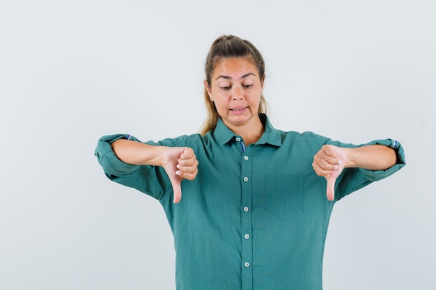 Mulher jovem com blusa verde mostrando os polegares para baixo e parecendo descontente