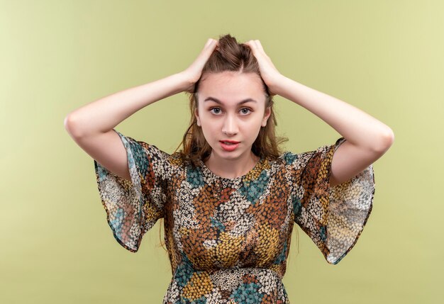 Foto grátis mulher jovem com cabelo comprido usando um vestido colorido estressada e frustrada tocando a cabeça em pé sobre a parede verde