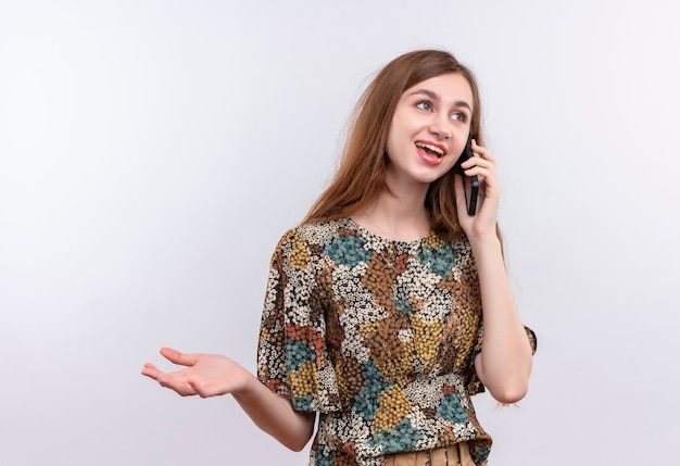 Mulher jovem com cabelo comprido usando um vestido colorido sorrindo enquanto fala no celular em pé sobre uma parede branca