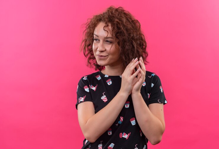 Foto grátis mulher jovem com cabelo curto e encaracolado com as palmas das mãos juntas olhando para o lado esperando em pé sobre a parede rosa
