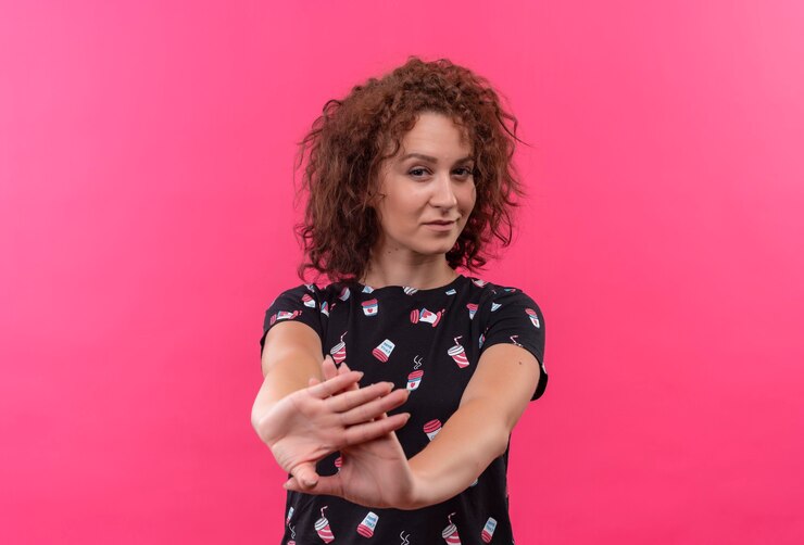 Foto grátis mulher jovem com cabelo curto e encaracolado com os braços abertos, fazendo gesto de defesa, parecendo confiante em pé sobre uma parede rosa