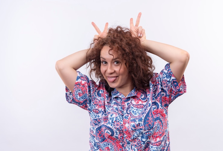 Foto grátis mulher jovem com cabelo curto e encaracolado com uma camisa colorida se divertindo, mostrando a língua e imitando chifres na cabeça com os dedos sobre a parede branca