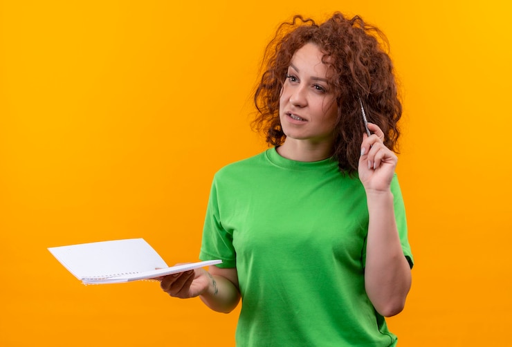 Foto grátis mulher jovem com cabelo curto e encaracolado com uma camiseta verde segurando um caderno e uma caneta olhando para o lado com uma expressão pensativa no rosto em pé